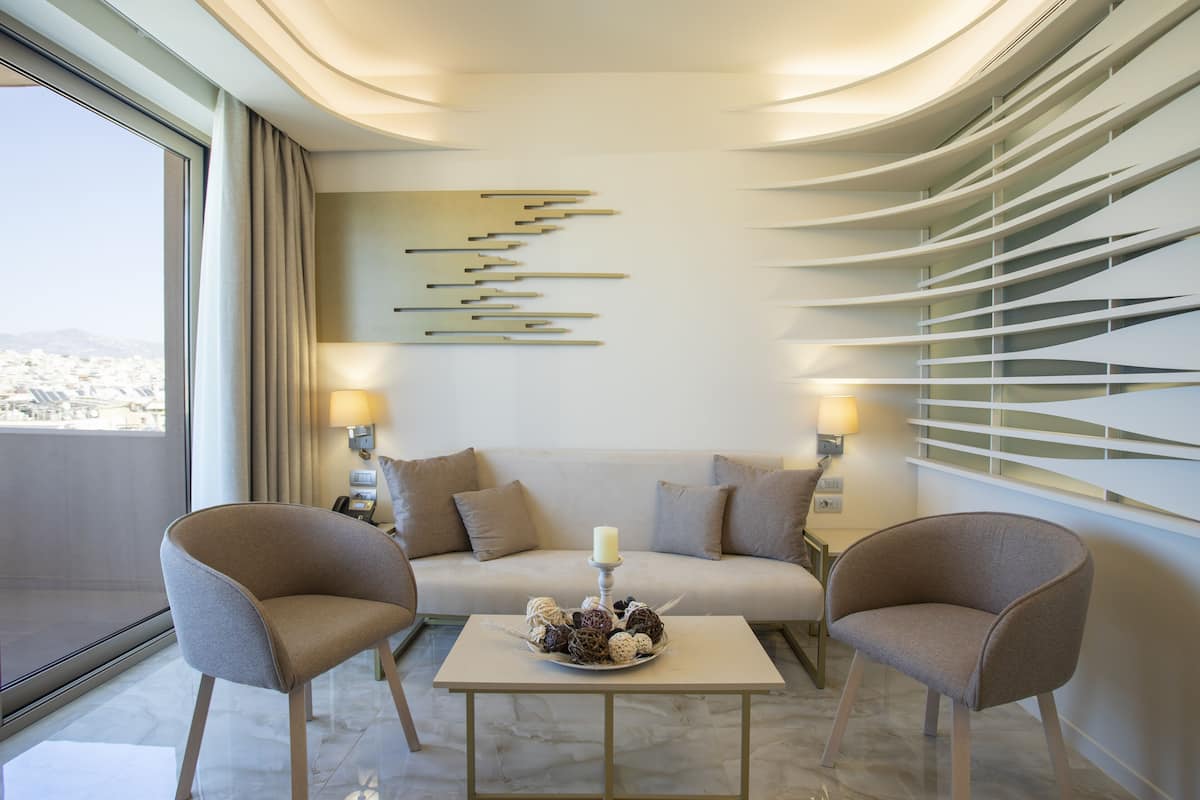 A modern hotel room with a sofa, two armchairs, a table with decorative items, and a city view through large windows.