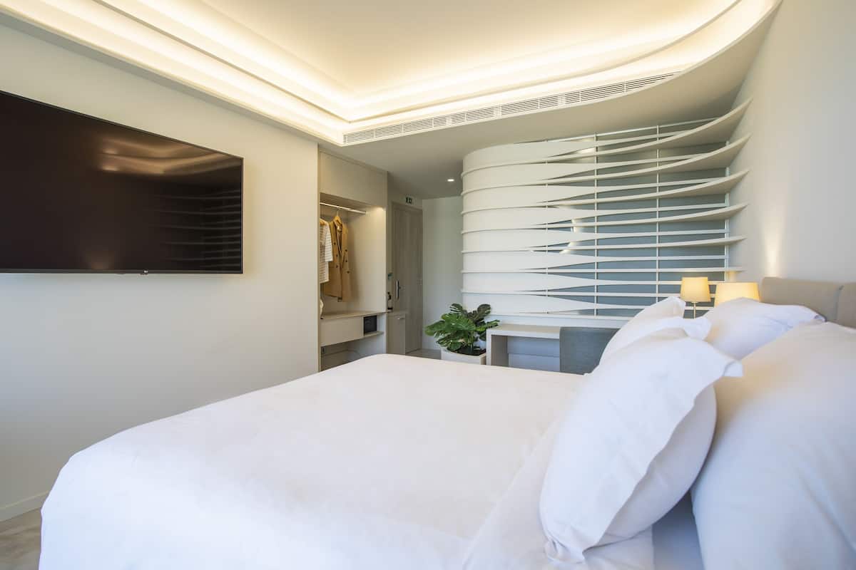 A modern hotel room with a large bed, a flat-screen TV mounted on the wall, a curved ceiling design, and a small bedside table with a lamp.