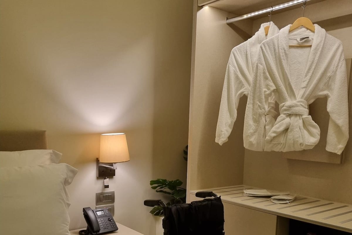 A hotel room with a bed, a bedside table with a lamp, a phone, a wheelchair, a robe, and a mirror.