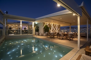 Un área de piscina moderna al aire libre con cerramiento de vidrio, asientos y vistas de la ciudad.