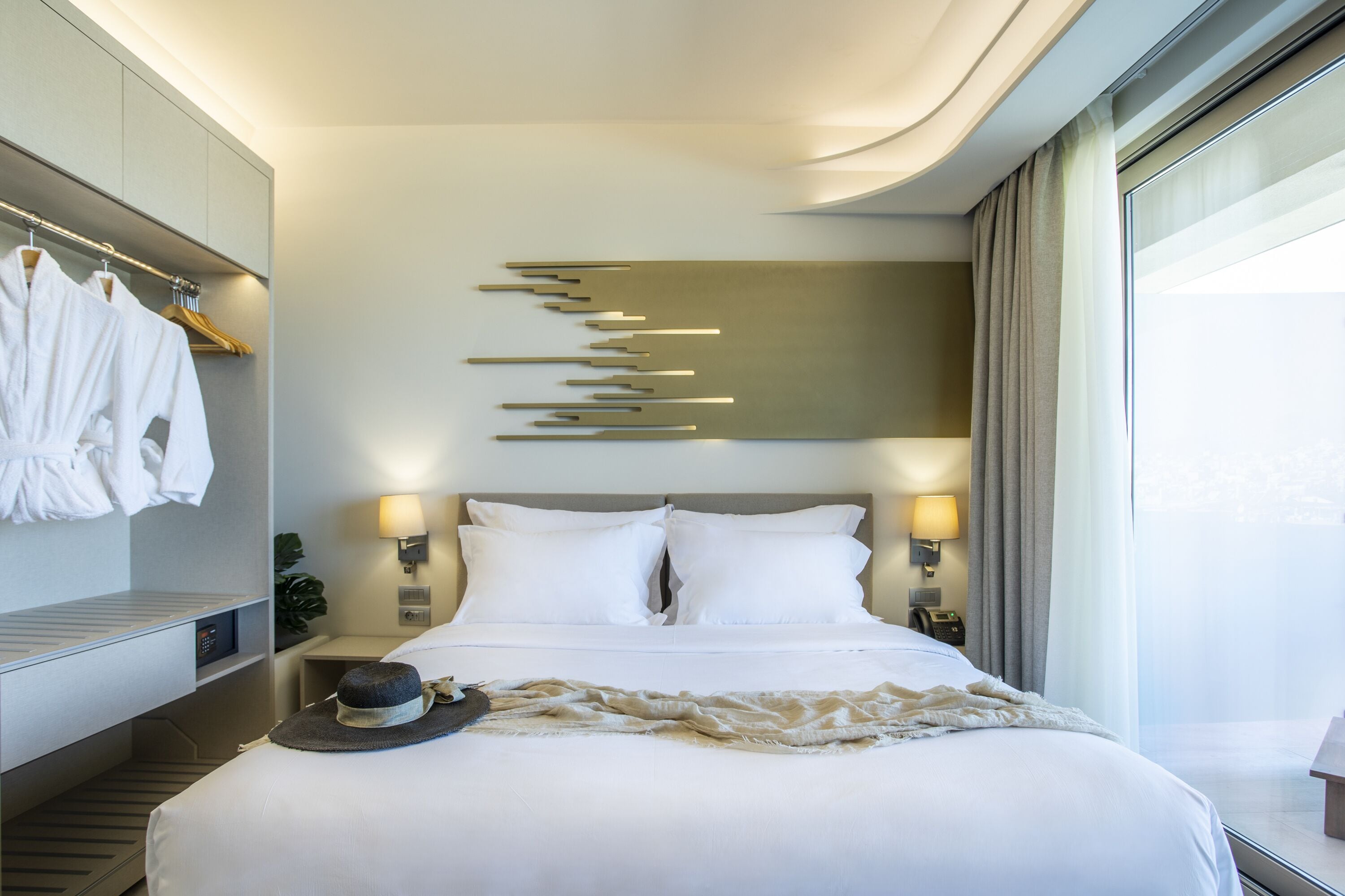 Une chambre d’hôtel moderne avec un lit bien fait, une table de chevet avec une lampe, un porte-peignoir fixé au mur et une grande fenêtre avec des rideaux.