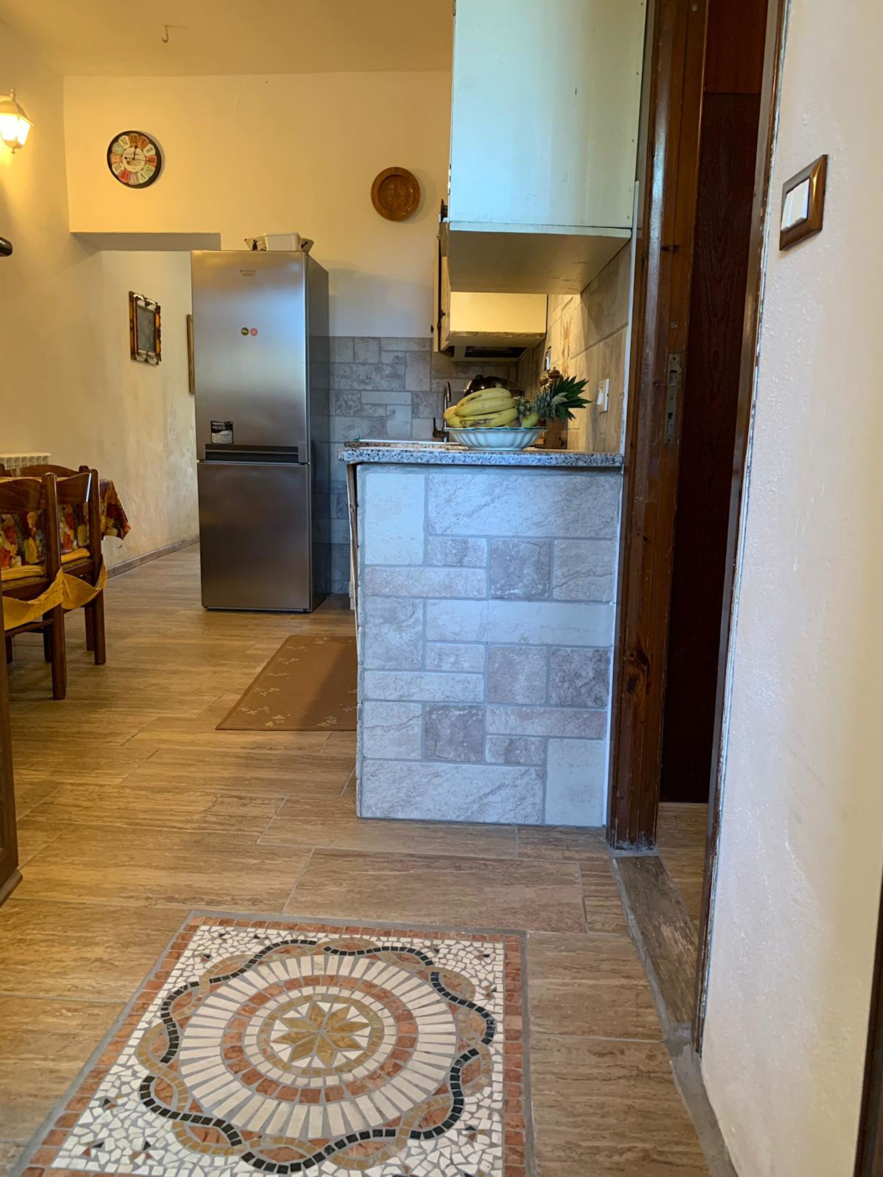 Eine Küche mit Kühlschrank, Spüle und einer Arbeitsfläche mit Früchten. Auf dem Boden liegt ein Teppich mit Muster.