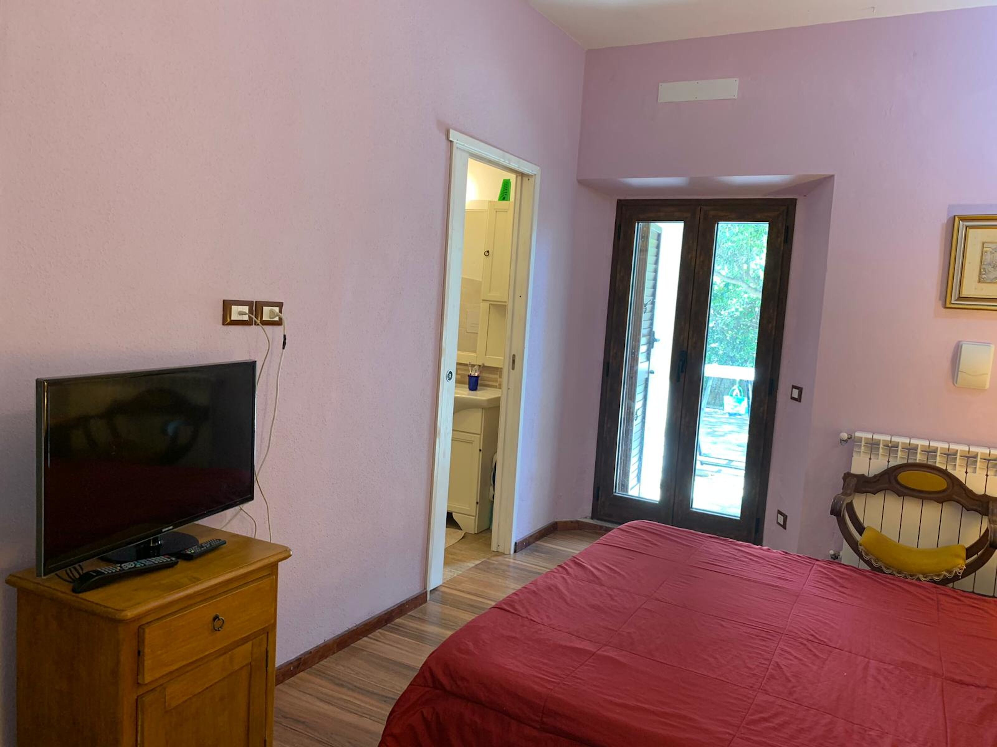 Un dormitorio con una televisión sobre un tocador de madera, una cama con una colcha roja, un cabecero de madera y una puerta que da a un baño.