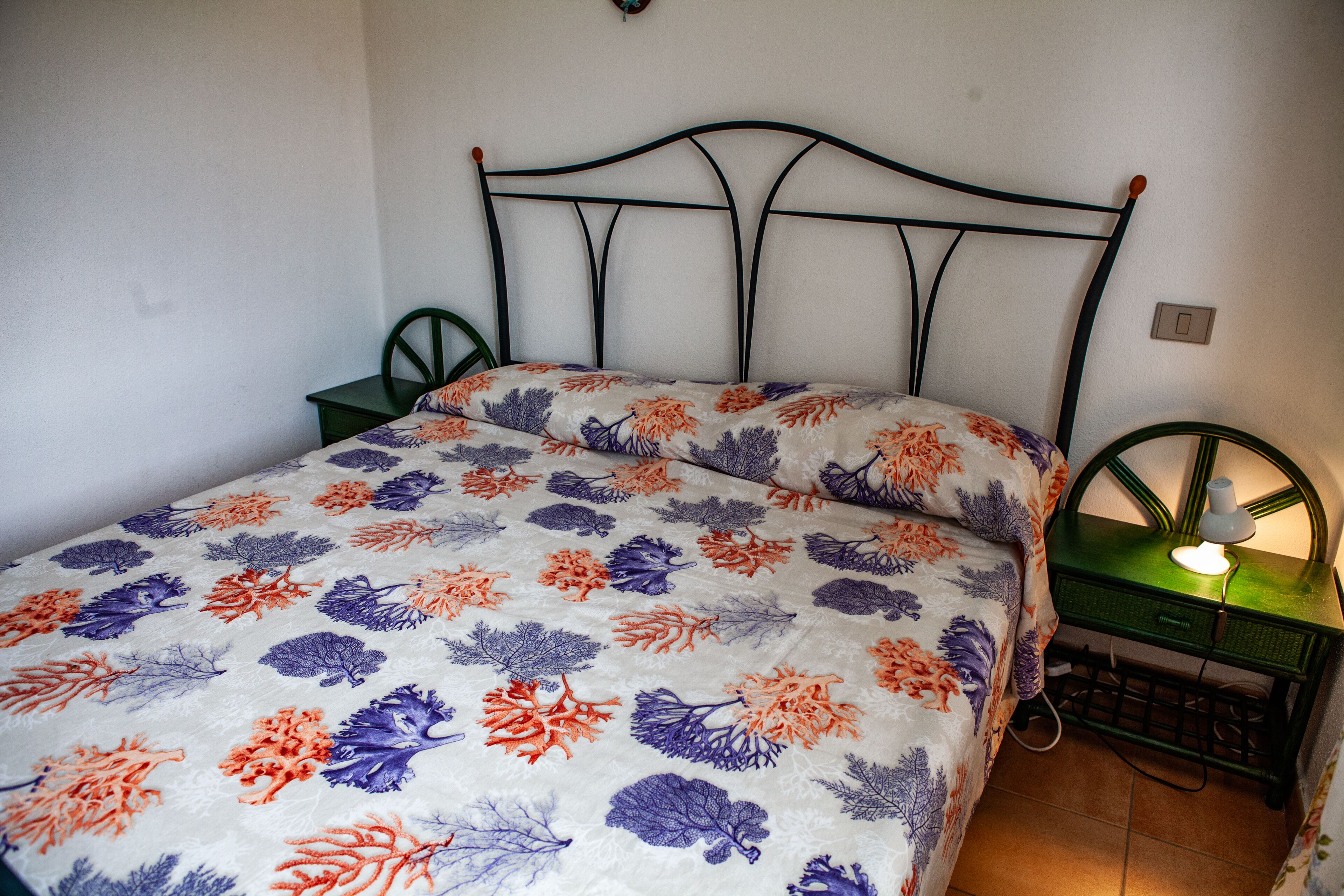 A bed with a floral bedspread, a green nightstand with a lamp, and a chair.
