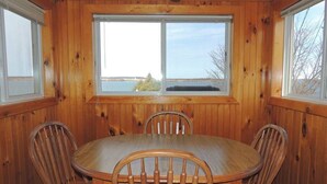 A wooden-paneled room with a round table and four chairs, large windows with a view of the outdoors, and a ceiling fan. - Cottage #6 - 2 bedroom/ 1 bath (Cape Vincent)