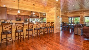 A spacious kitchen with wooden cabinets, a central island with stools, and a dining area with a sofa and chairs. - Free Tickets | 2 Hot Tubs, Arcade, Fireplace, Parking, Easy Access for Buses (Gatlinburg)