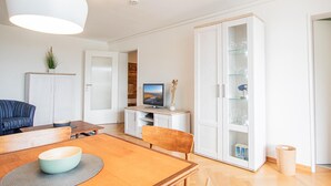 A modern living room with a wooden dining table, chairs, a TV on a stand, a cabinet with glass doors, and a white door. - App. Bagehorn 10 (Westerland)
