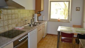 Een kleine keuken met een raam, een gootsteen, een kookplaat en een eethoek met een tafel en stoelen.