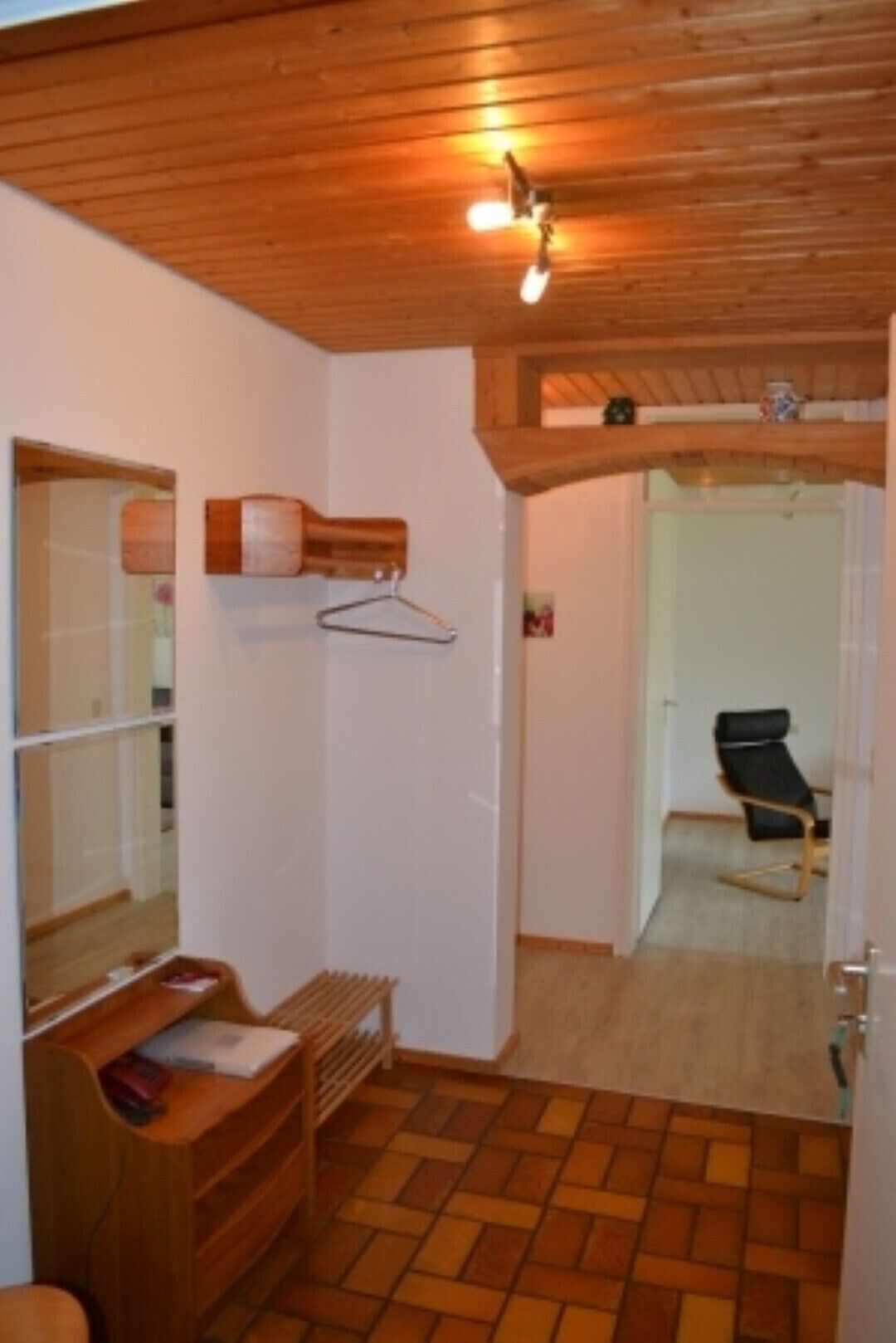 A room with a wooden ceiling, a hanging light fixture, a wooden floor, a chair, and a door leading to another room.