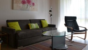Een woonkamer met een donkerbank, groene en gele kussens, een glazen salontafel en een zwarte fauteuil.