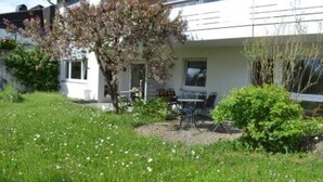 Een gebouw met twee verdiepingen, met een patio voorzien van een tafel en stoelen, omgeven door groen en een kleine boom met roze bloesems.