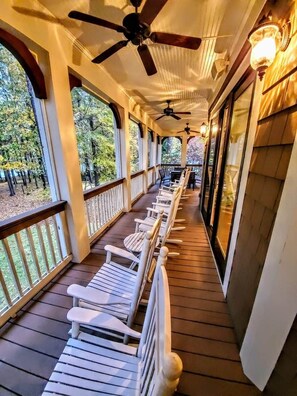 A covered porch with wooden flooring, white rocking chairs, ceiling fans, and large windows offering a view of trees.