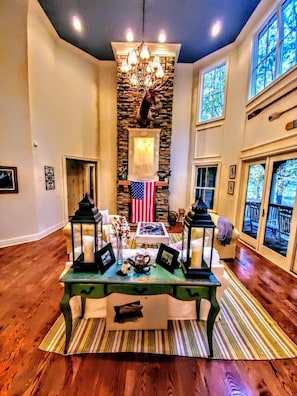 A spacious living room with a stone fireplace, a chandelier, and a view of the outdoors through large windows.