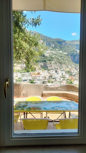 A view through a window of a terrace with yellow chairs and a table, overlooking a hillside with buildings and greenery.