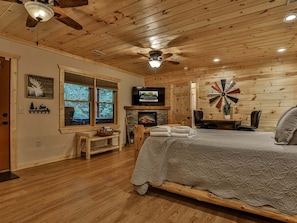A cozy log cabin bedroom with a bed, a fireplace, a television, and a large window.