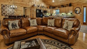 A cozy living room with a brown leather sectional sofa, wooden ceiling, and a kitchen area in the background. - ​​​​​​​Jagged Owl by Mountain Oasis | Dog-Friendly Cabin in Coosawattee River Resort (Ellijay)