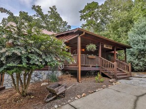 A wooden cabin with a porch, surrounded by trees and a stone wall.