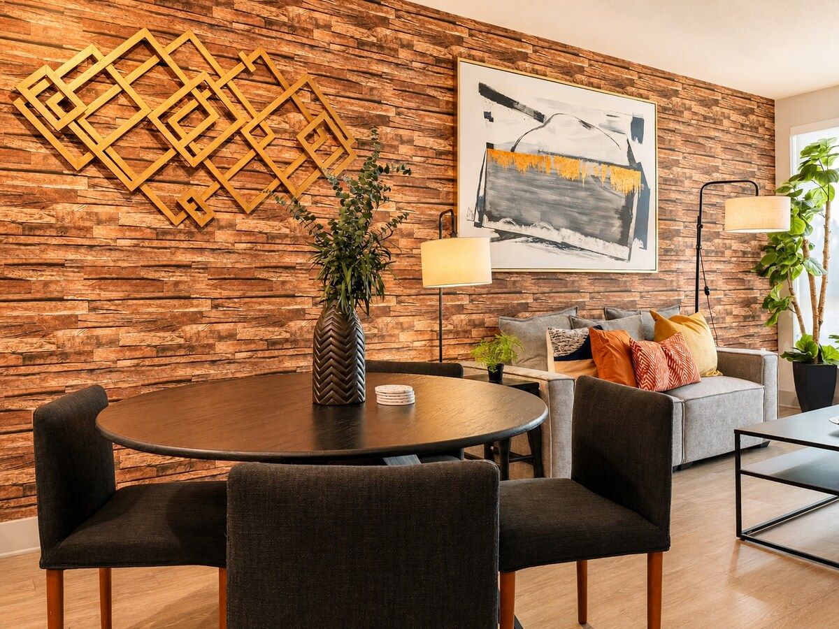 A modern living room with a round dining table, dark upholstered chairs, a grey sofa with cushions, a large abstract painting, a geometric wall decoration, and a potted plant.