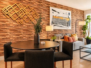 Un salon moderne avec une table à manger ronde, des chaises à revêtement sombre, un canapé gris avec des coussins, une grande peinture abstraite, un élément décoratif géométrique sur le mur et une plante en pot.