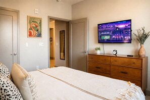 A bedroom with a bed, a dresser, a television mounted on the wall, and a door leading to another room. - Resort-Style Condo 3 Miles to Disney (Fullerton)