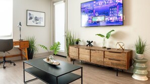 A modern living room with a flat-screen TV, wooden cabinet, desk, chair, and potted plants. - Resort-Style Condo 3 Miles to Disney (Fullerton)