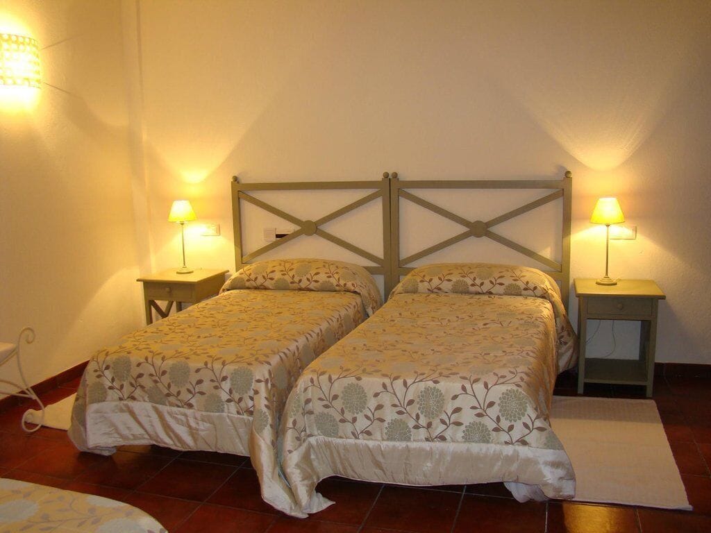 Una habitación con dos camas individuales, cada una con una mesita de noche y una lámpara. El suelo es de baldosas y la pared, lisa.