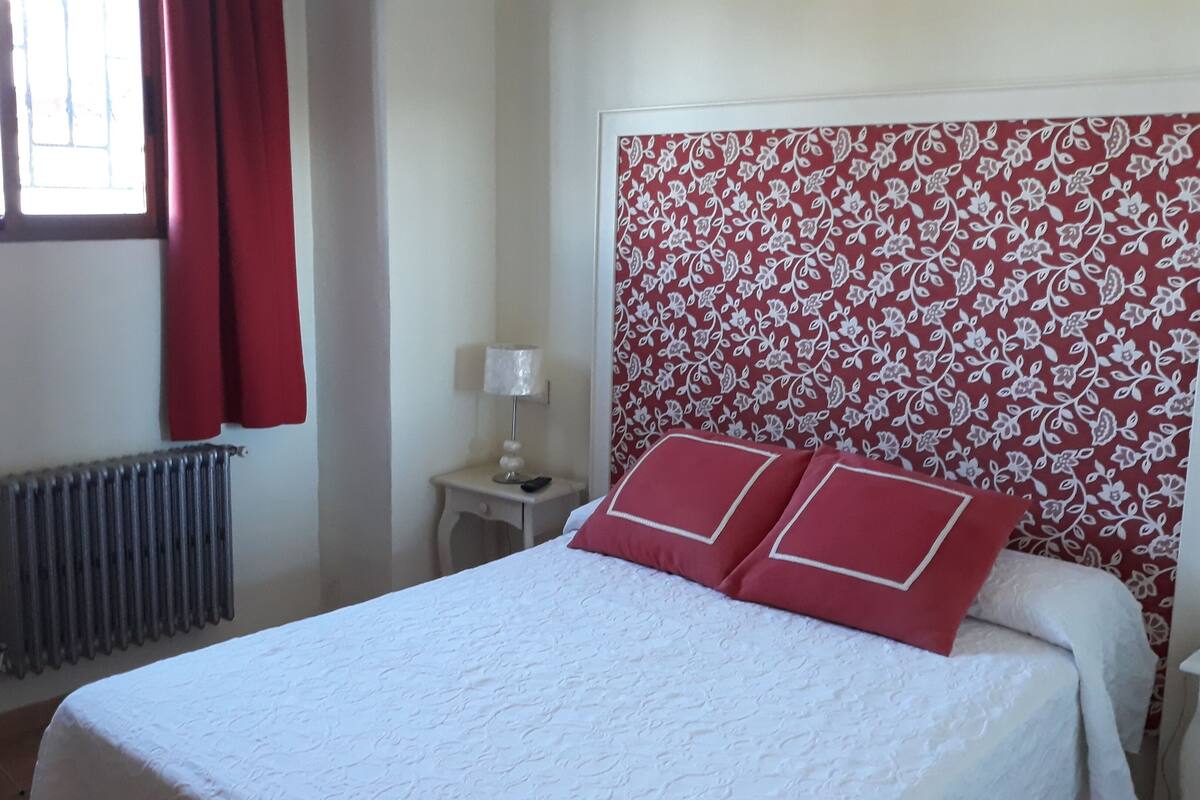 A hotel room with a bed, a radiator, a nightstand with a lamp, and a window with red curtains.