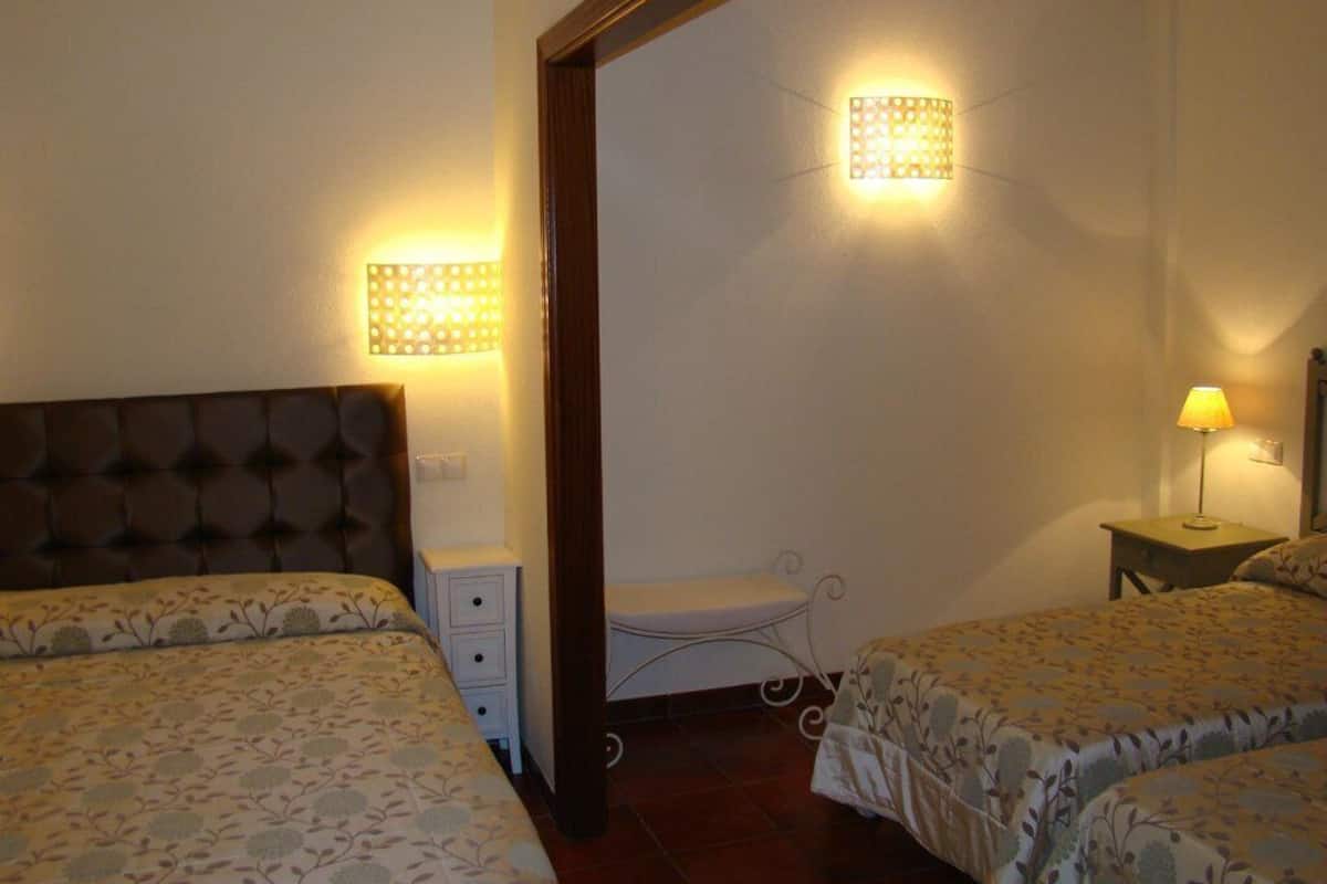 A bedroom with two beds, a nightstand, a small table with a lamp, and a wall-mounted light fixture.