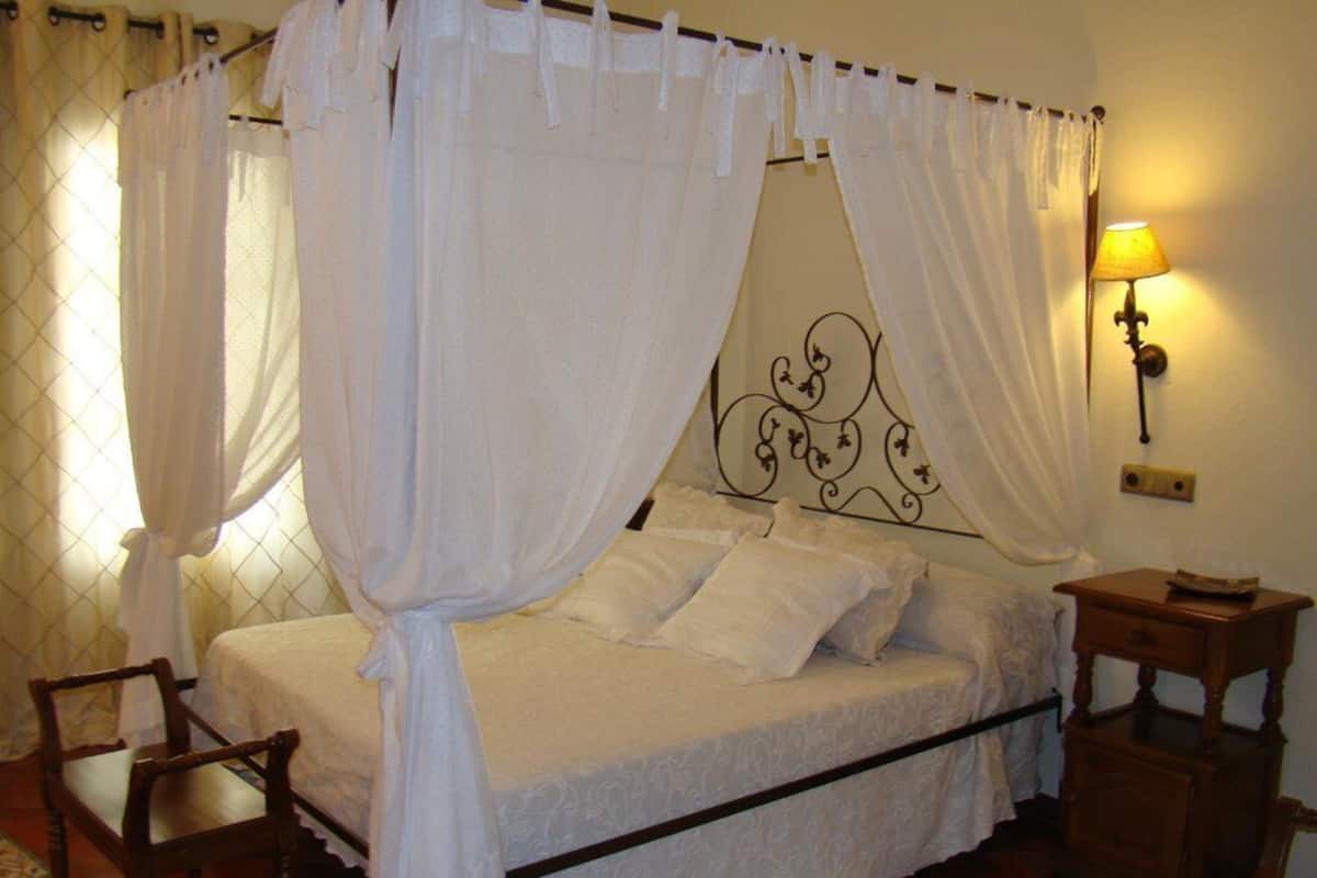 A bedroom with a four-poster bed, white bedding, a wooden nightstand, and a wall-mounted lamp.