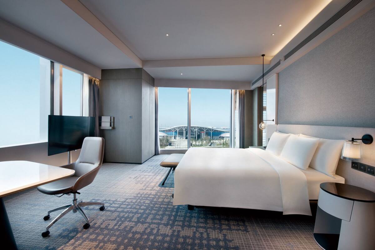 A modern hotel room with a large bed, a desk, a chair, and a TV. The room has large windows with a view of a bridge.