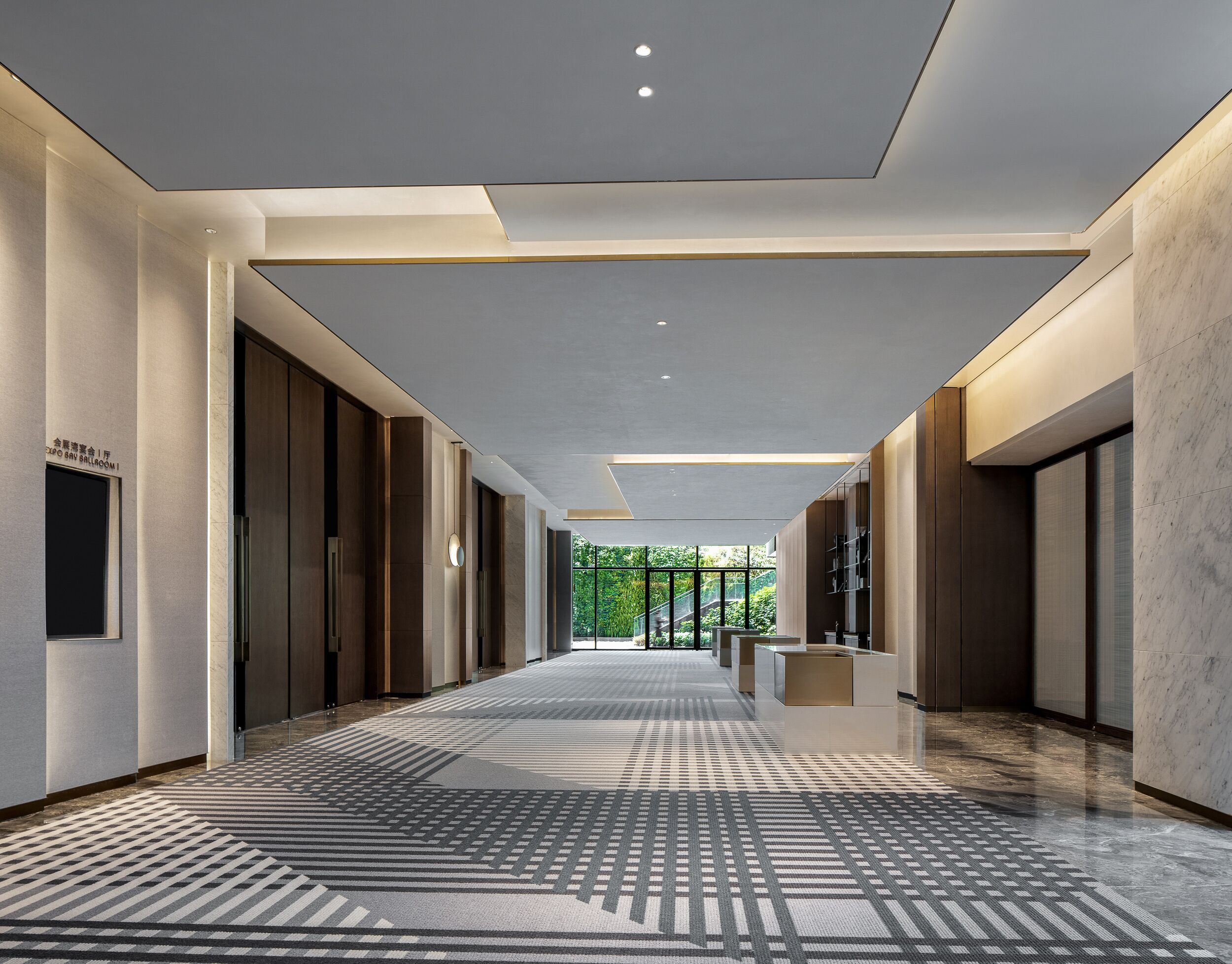Een moderne hotelgang met een kamerbreed tapijt, inbouwverlichting en uitzicht op groen.