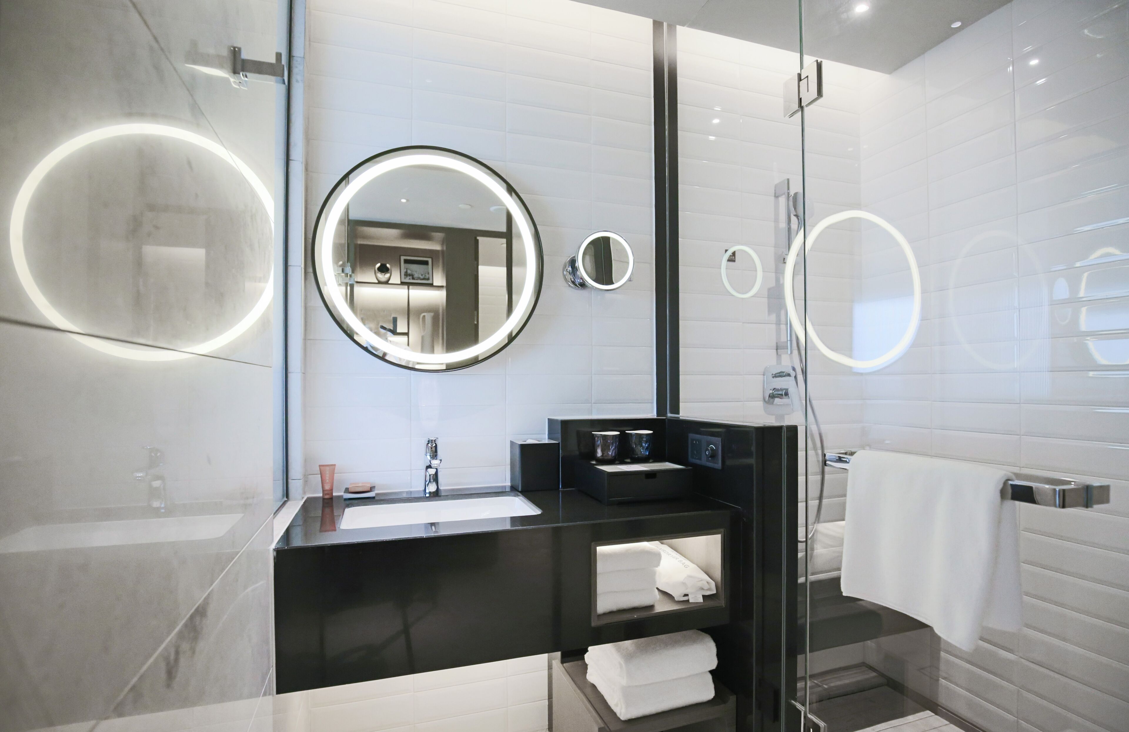 Un baño moderno con cabina de ducha de vidrio, un espejo redondo con iluminación integrada, encimera negra y toallero blanco.