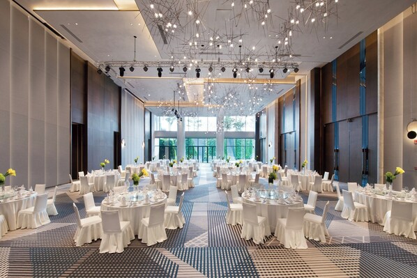Une vaste salle de banquet avec des tables rondes, des nappes blanches et des chaises, agrémentée d’un tapis à motifs et d’un lustre moderne.