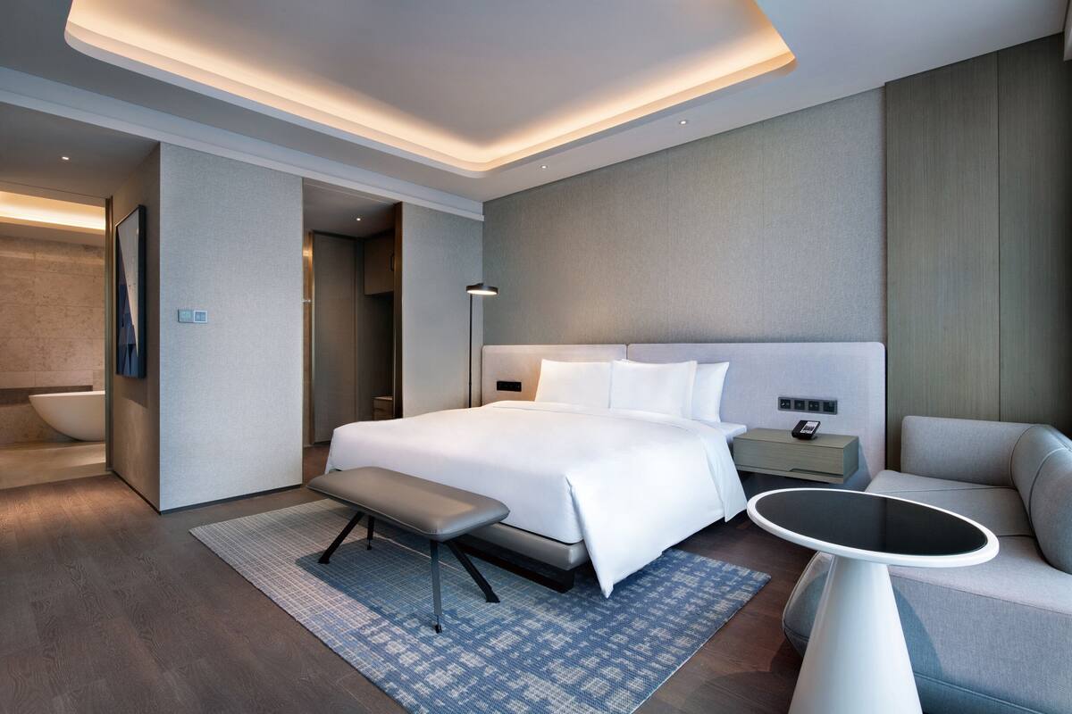 Une chambre d’hôtel moderne dotée d’un grand lit, de tables de chevet, d’un banc, d’une table ronde et d’une salle de bain avec baignoire.