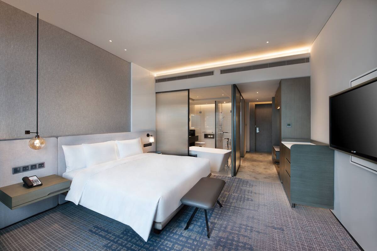 Une chambre d’hôtel moderne dotée d’un grand lit, de tables de chevet, d’un bureau, d’un téléviseur à écran plat et d’une salle de bain visible à travers une paroi en verre.