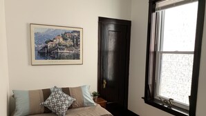 A bedroom with a bed, pillows, a framed picture, a dark wooden door, and a window with a view of buildings.
