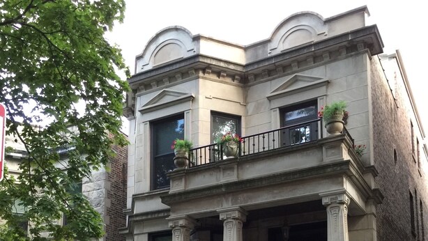 A two-story building with a classical architectural style, featuring a balcony with potted plants, a gated entrance, and a visible address number.