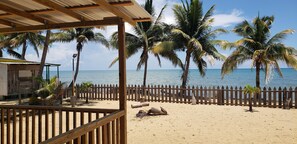 A beach with a wooden fence, palm trees, and a wooden structure with a roof. - Pal's on the Beach - A Gold Standard Hotel (Dangriga)