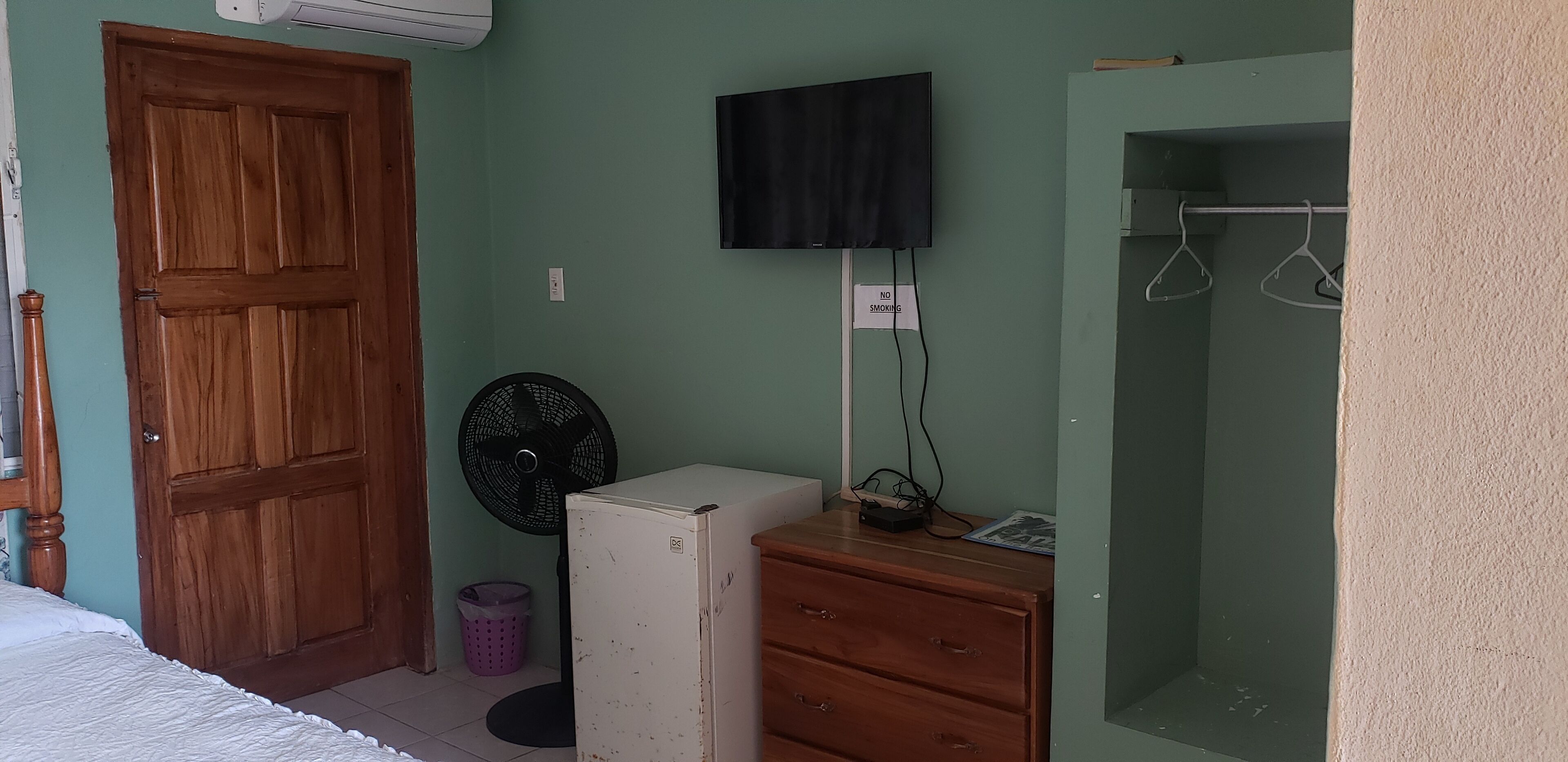 A room with a wooden door, a white refrigerator, a fan, a television, and a small wooden dresser.