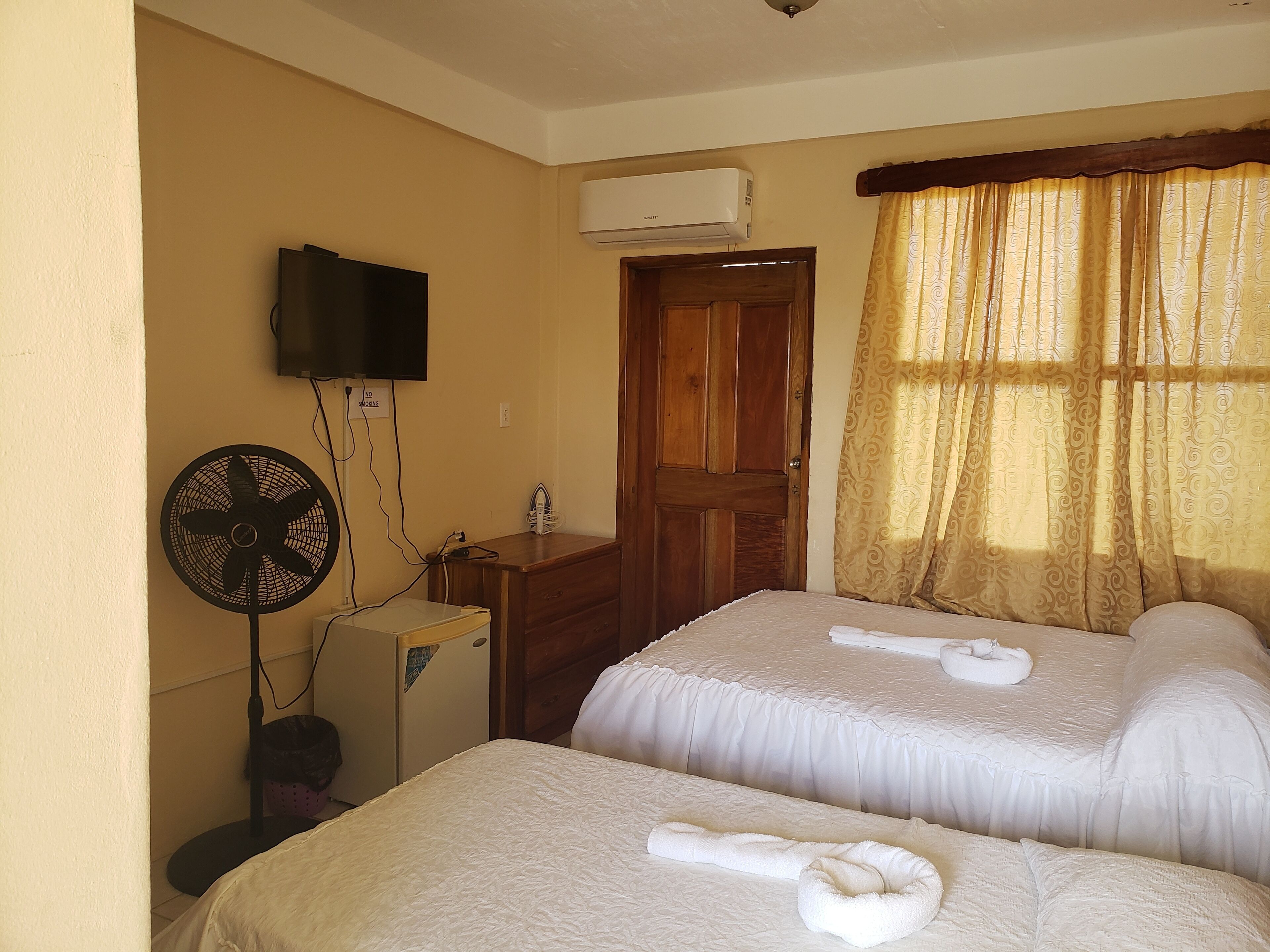 A hotel room with two beds, a TV, an air conditioner, a fan, and a wooden door.