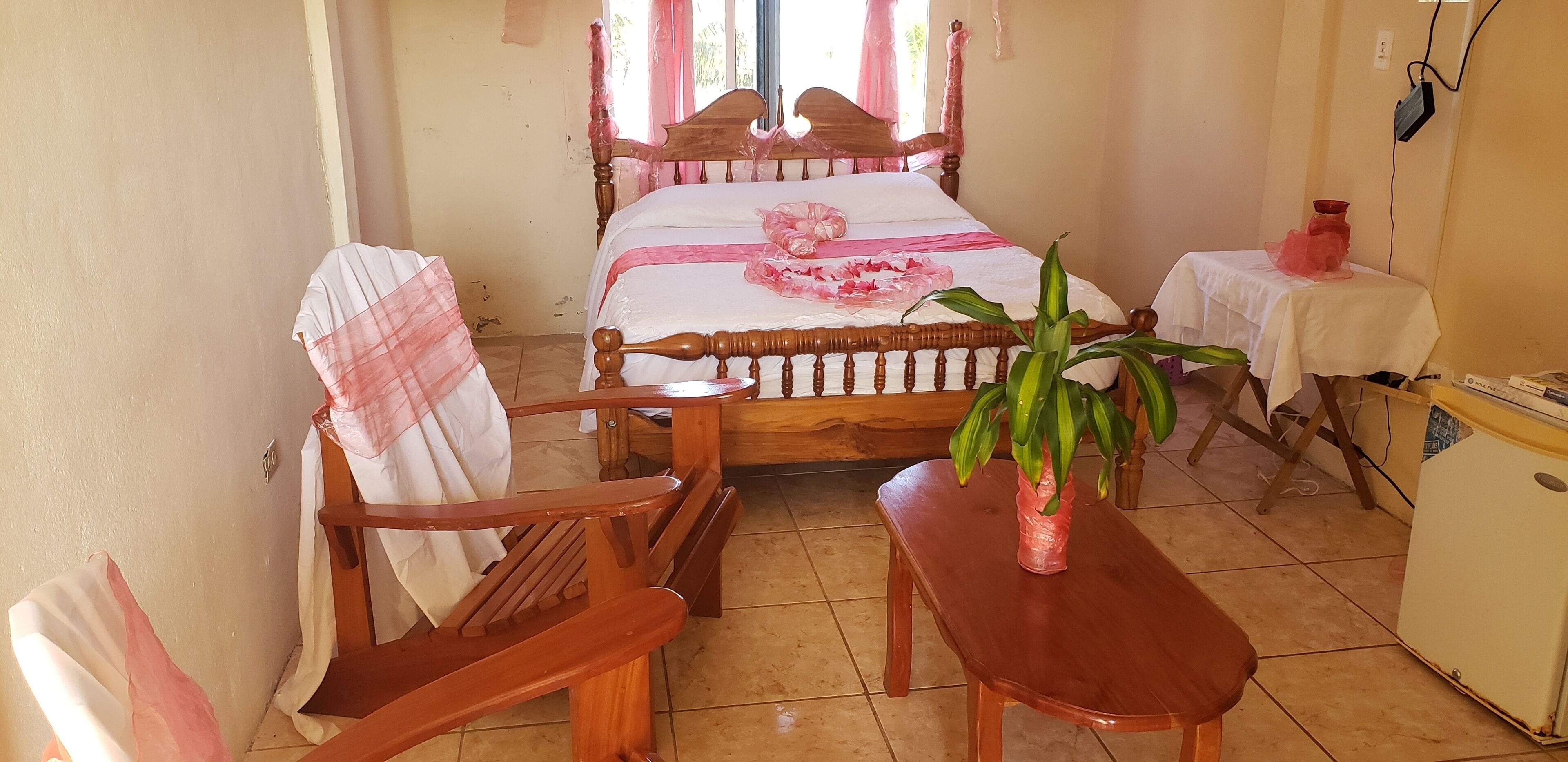 A bedroom with a wooden bed, a chair, a small table, a potted plant, and a bedside table with a lamp.