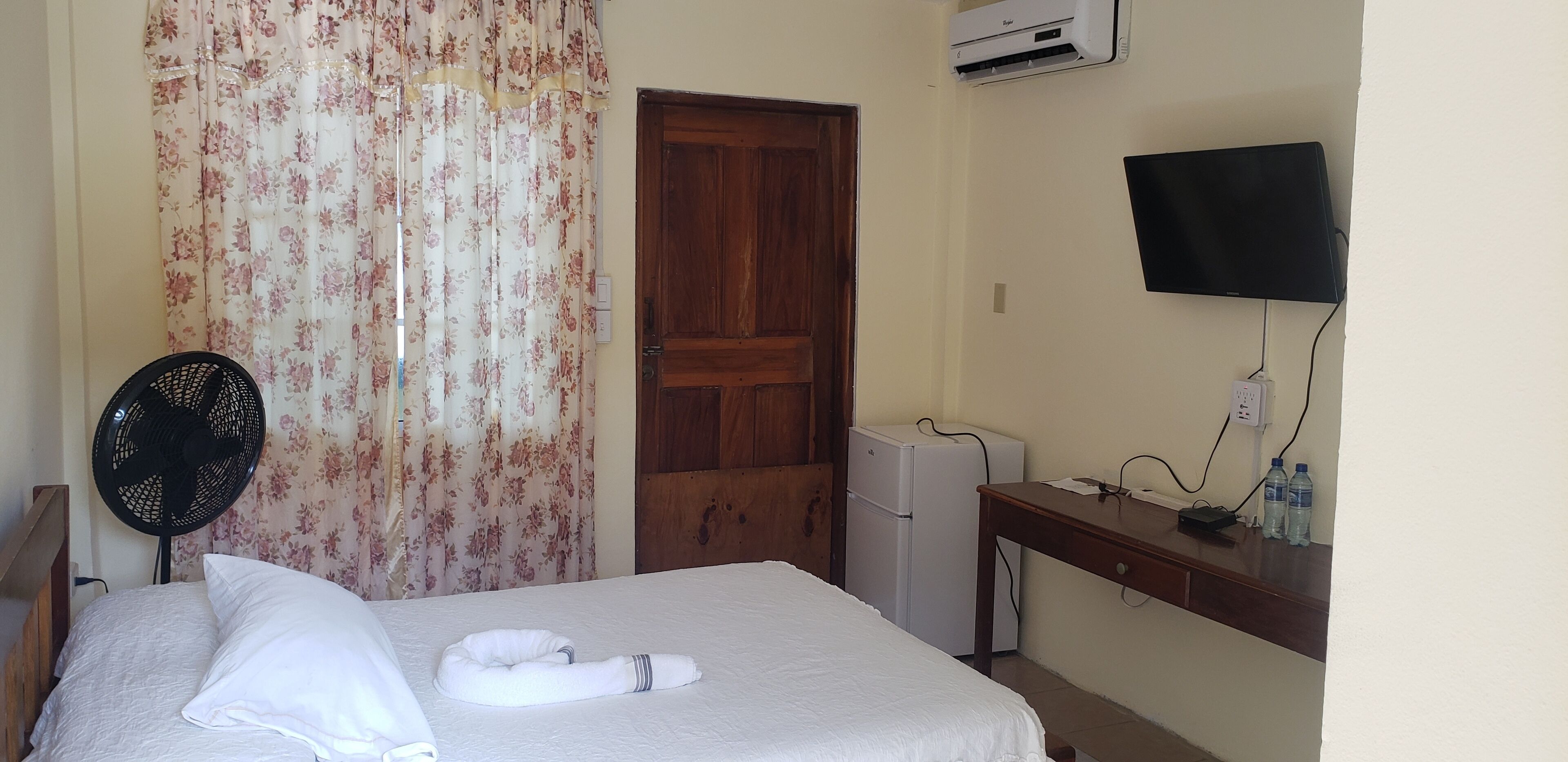 Une chambre d’hôtel équipée d’un lit, d’un ventilateur, d’une télévision, d’un mini-réfrigérateur et d’un distributeur d’eau.