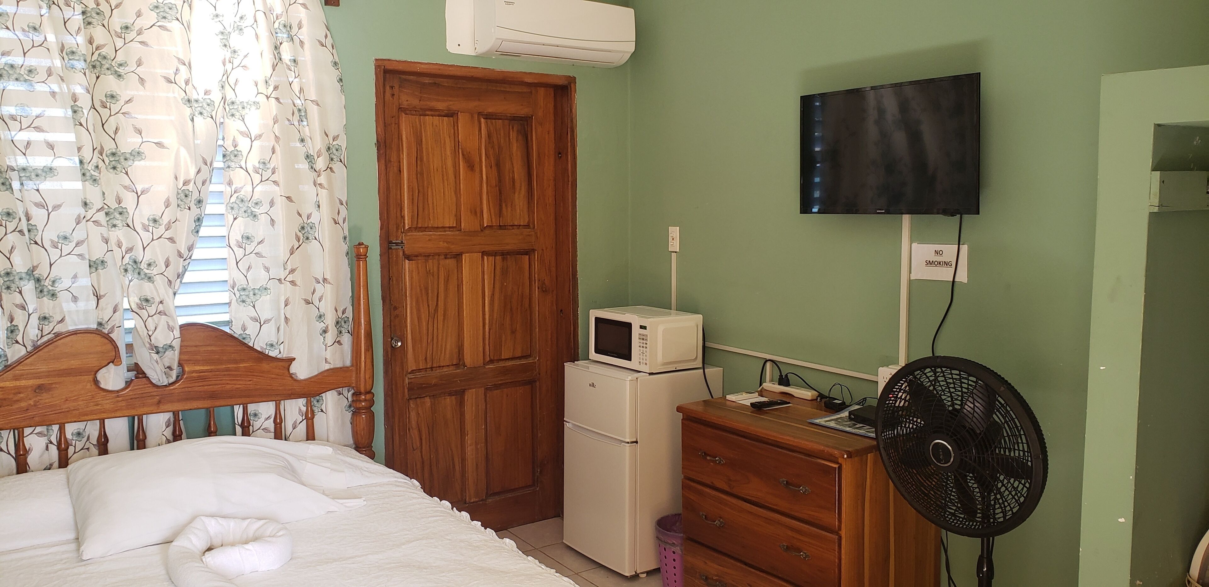 A bedroom with a wooden bed, a white mattress, a wooden dresser, a black fan, a microwave, and a television on the wall.