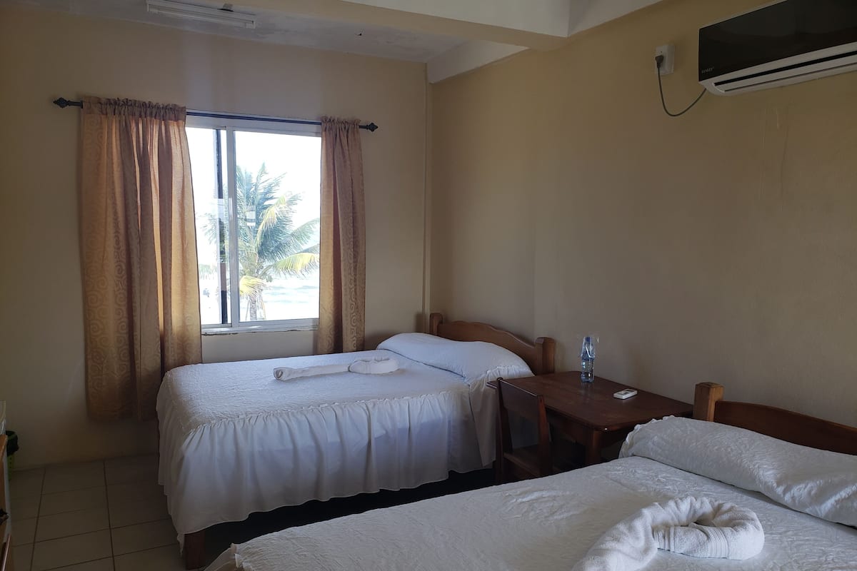 Habitación de hotel con dos camas, una mesita, una silla, una botella de agua y un televisor fijado a la pared.