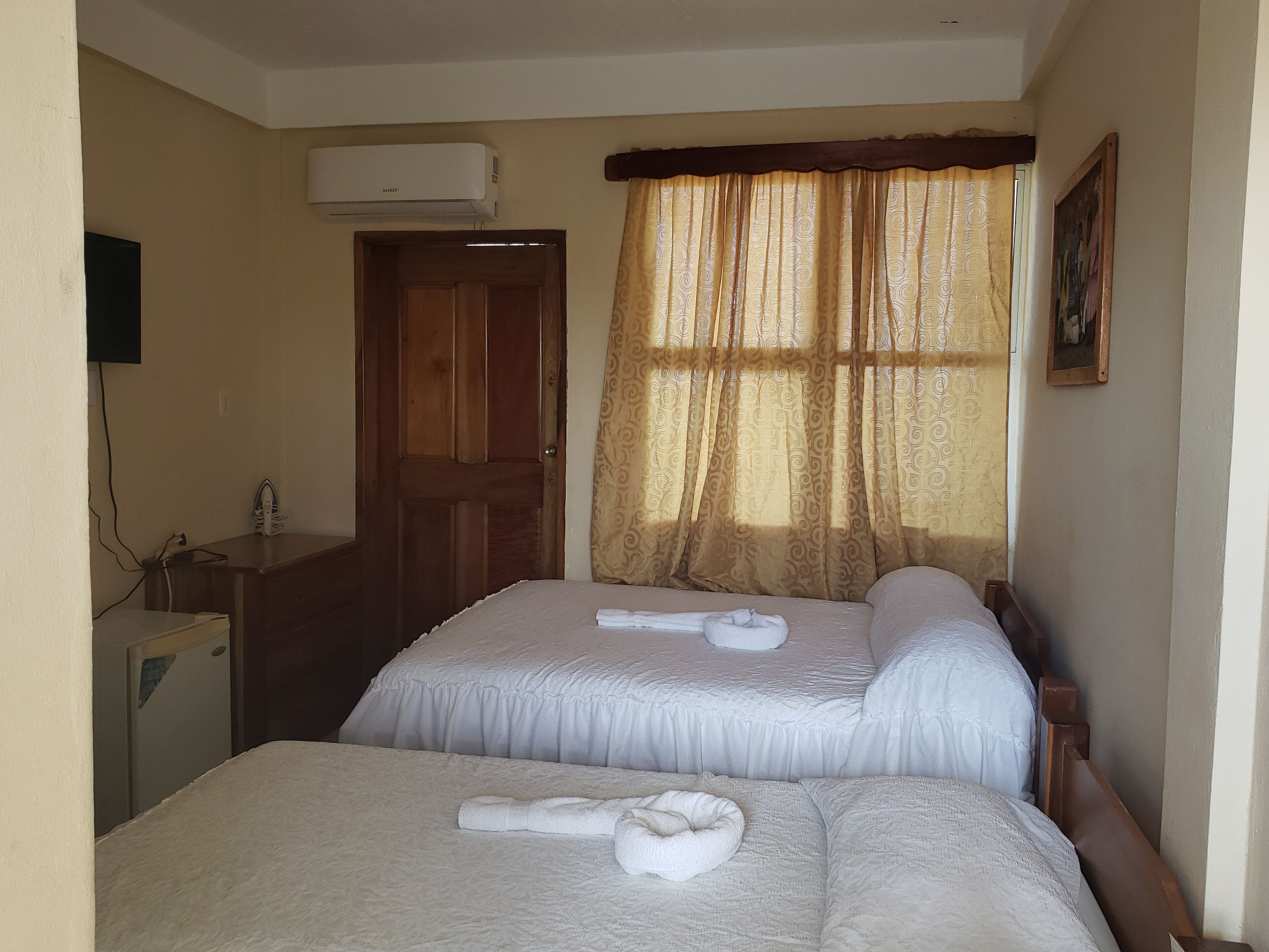 A hotel room with two beds, a wooden wardrobe, a desk with a chair, an air conditioning unit, and a painting on the wall.