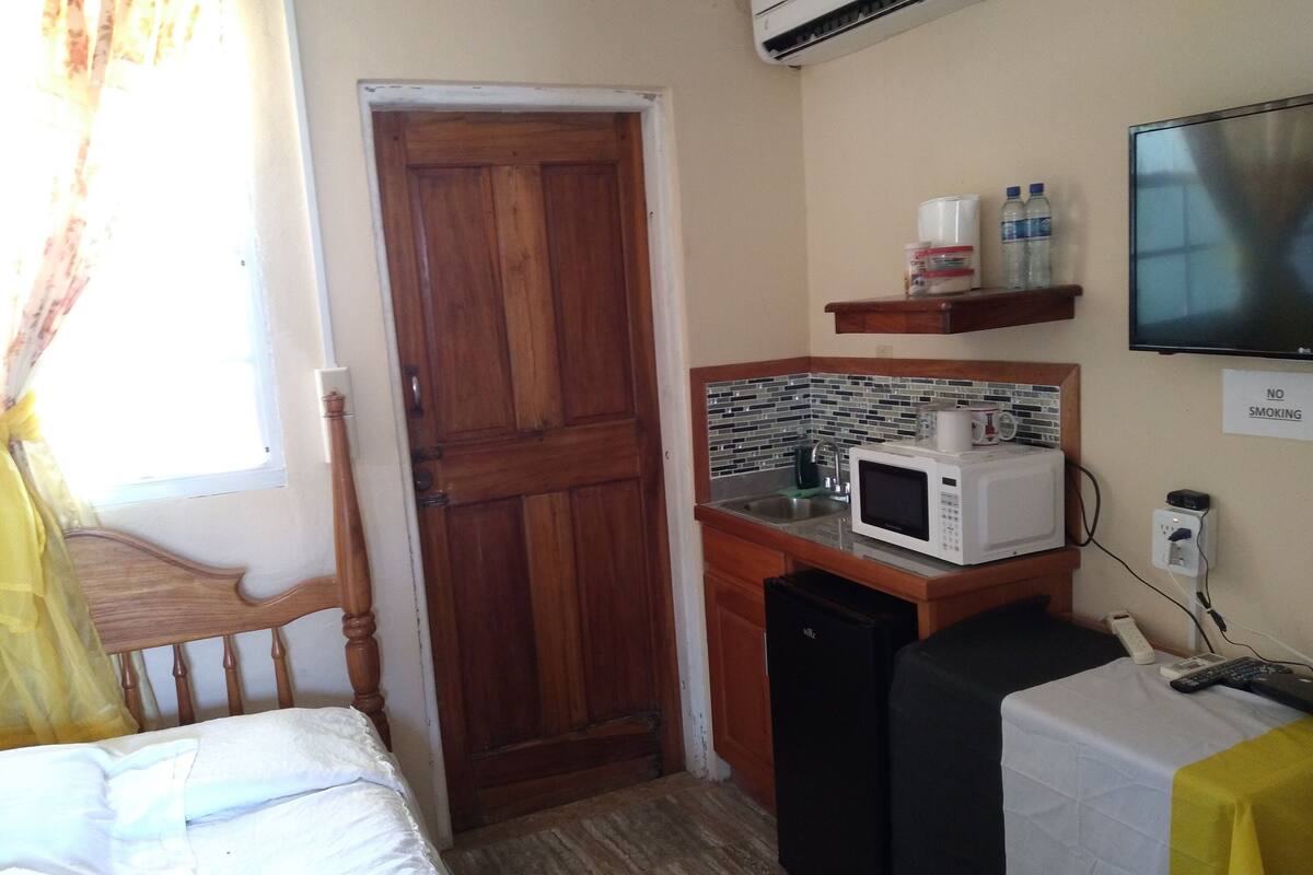 Habitación de hotel pequeña con puerta de madera, una kitchenette con microondas, lavamanos y televisión en la pared.