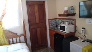 Habitación de hotel pequeña con puerta de madera, una kitchenette con microondas, lavamanos y televisión en la pared.
