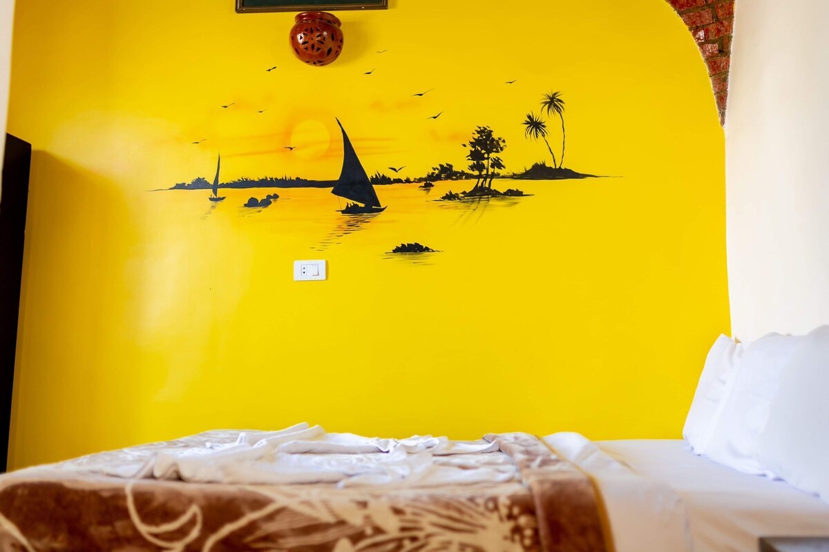 A room with a bed, a wall with a yellow mural of a sunset and sailboats, and a small wall-mounted light fixture.