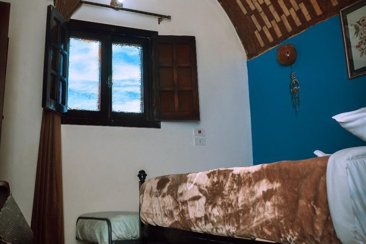 A room with a bed, a window with curtains, a wall-mounted heater, and a small table with a chair.