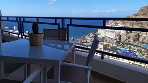 A balcony with a table and chairs, a potted plant, and a view of a coastal resort area with buildings and a pool. - Amazing ocean view apartment in Taurito, Mogán (Mogan)